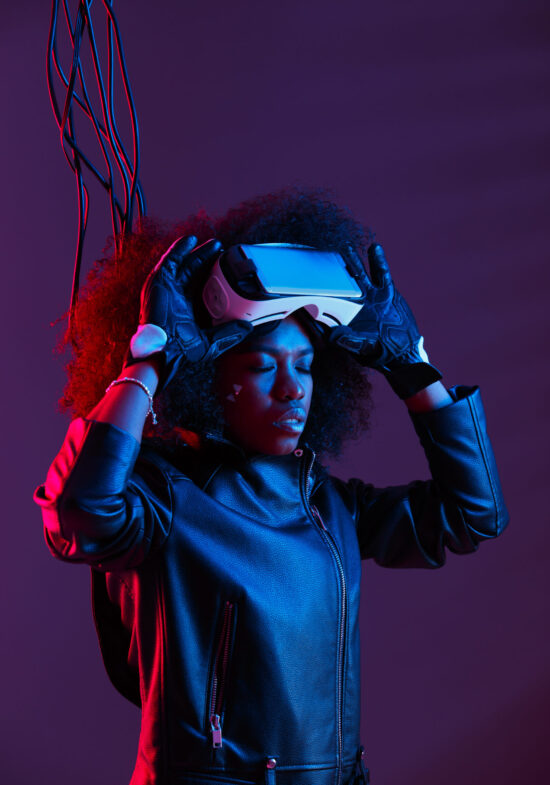 Curly dark haired girl dressed in a black leather jacket and gloves is wearing  the virtual reality glasses on her head in the dark studio with neon light . Cyberpunk character